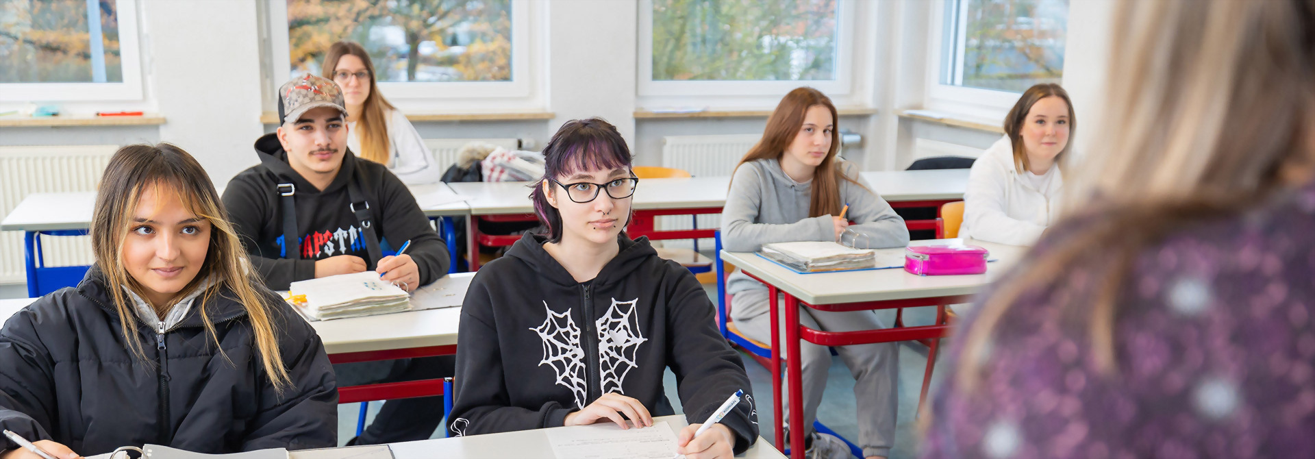 Schüler sitzen in einem hellen Klassenraum an Tischen und schreiben in Hefte.
