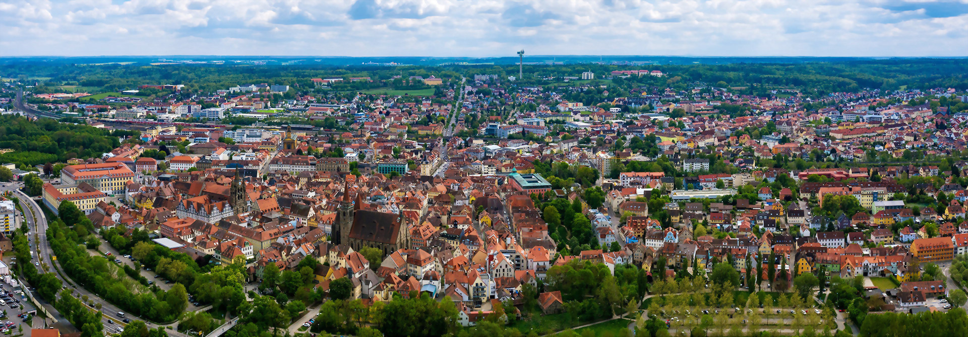 Luftaufnahme der Stadt Ansbach mit dicht bebautem Stadtzentrum und umliegender Landschaft unter blauem Himmel mit Wolken.