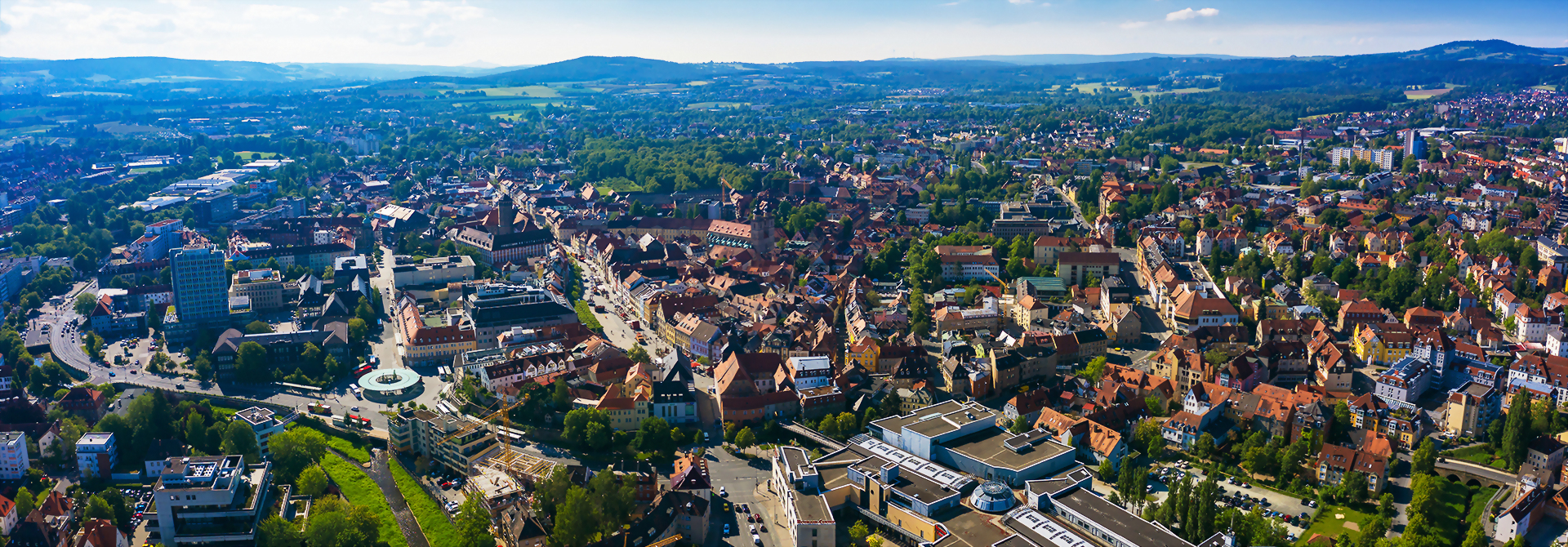 Panoramablick auf die Stadt Bayreuth mit zahlreichen Gebäuden, grünen Flächen und Hügeln im Hintergrund.