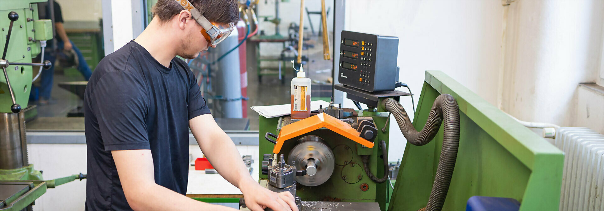 Mann in schwarzem T-Shirt arbeitet an einer grünen Maschine in einer Werkstatt mit Flasche und Steuergerät im Hintergrund.
