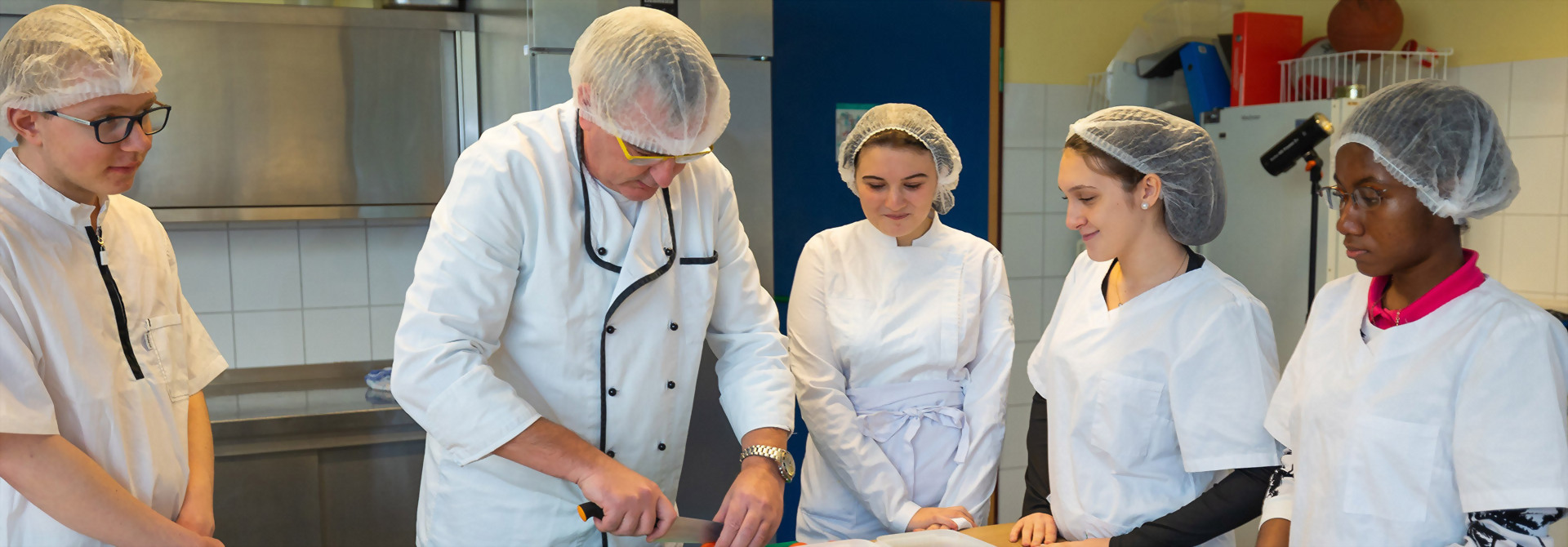 Mehrere Personen in Kochkleidung und Haarnetzen stehen um eine Arbeitsfläche in einer Küche, eine Person schneidet eine Tomate.