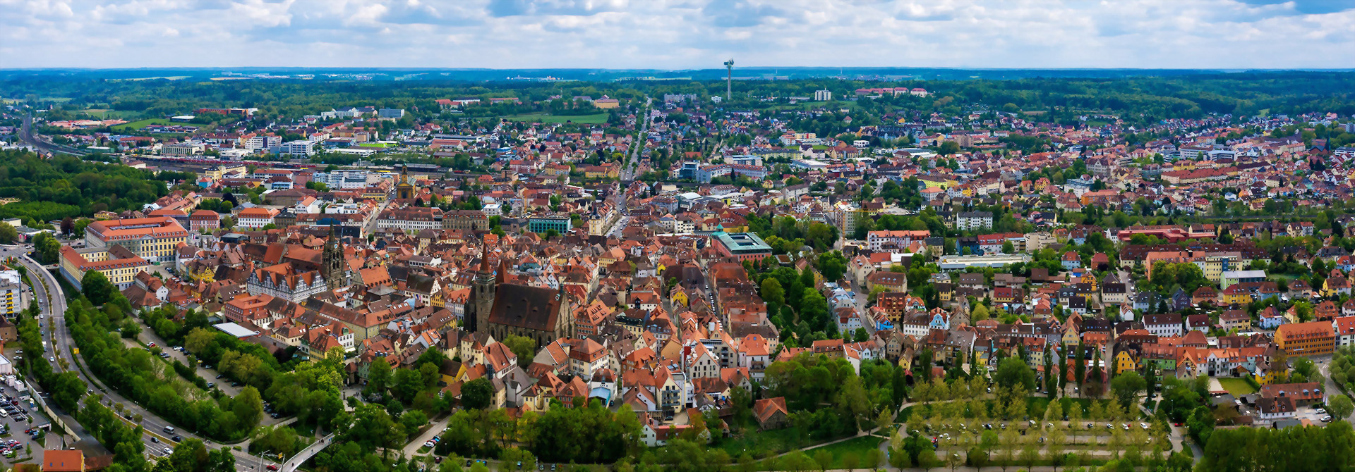 Luftaufnahme der Stadt Ansbach mit dicht bebautem Stadtzentrum und umliegender Landschaft unter blauem Himmel mit Wolken.
