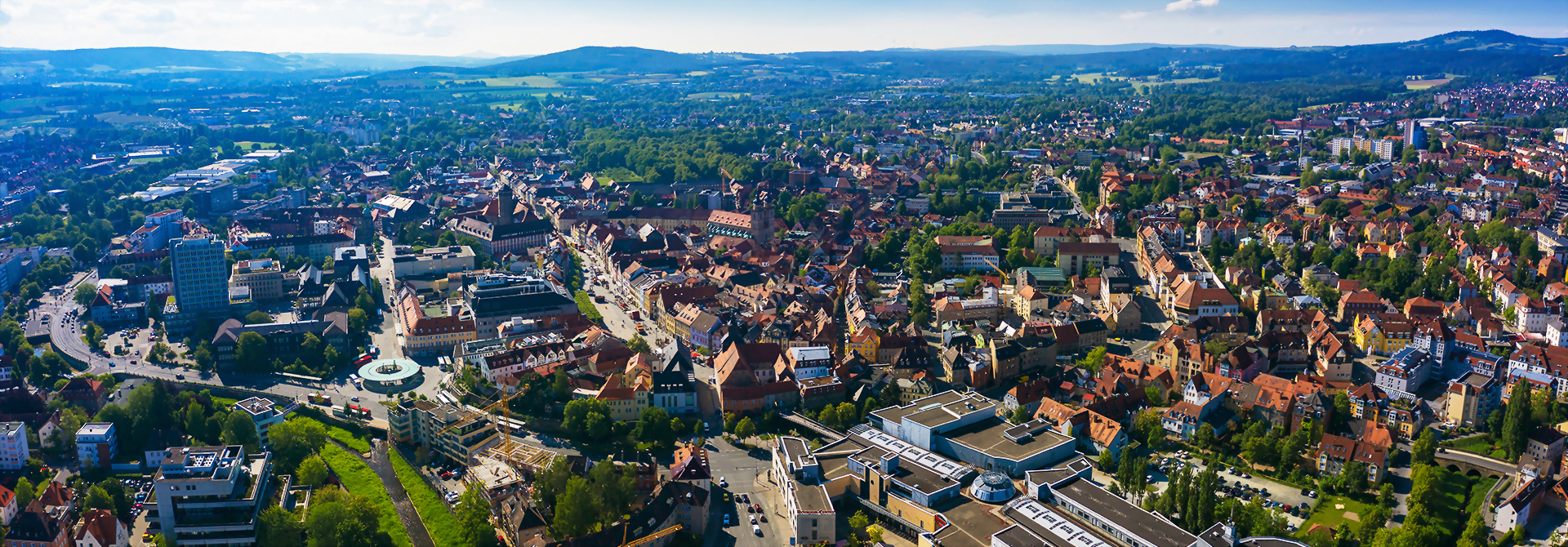 Panoramablick auf die Stadt Bayreuth mit zahlreichen Gebäuden, grünen Flächen und Hügeln im Hintergrund.