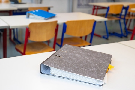 Ordner liegt auf einem weißen Tisch im Vordergrund, im Hintergrund mehrere leere Schulstühle und Tische in einem Klassenraum