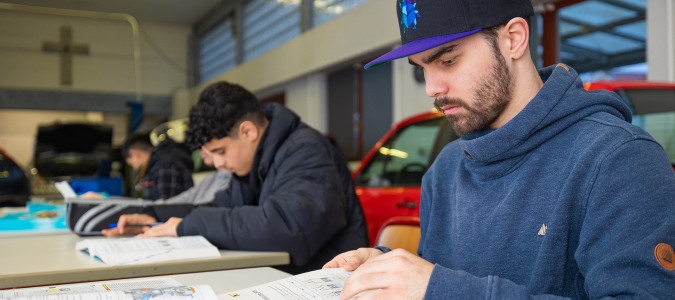 Mehrere Personen sitzen an Tischen und lesen oder schreiben in einem hellen Raum mit großen Fenstern und einem roten Auto im Hintergrund.