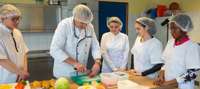 Mehrere Personen in Kochkleidung und Haarnetzen stehen um eine Arbeitsfläche in einer Küche, eine Person schneidet eine Tomate.