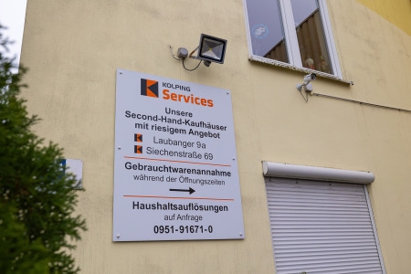 Schild an einer Hauswand mit Hinweisen zu Second-Hand-Kaufhäusern und Gebrauchtwarenannahme der Kolping Services inklusive Adressen und Telefonnummer.