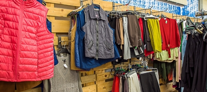 Mehrere Kleidungsstücke wie Jacken, Westen und Pullover hängen an einer Holzwand in einem Second-Hand-Laden.