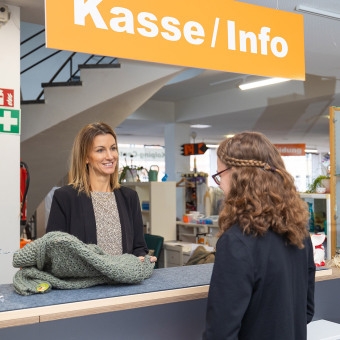 Zwei Frauen an einem Tresen unter einem Schild mit der Aufschrift 'Kasse / Info' in einem Second-Hand-Laden.