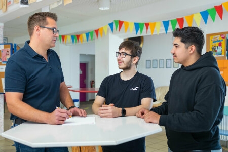 Drei Personen stehen an einem weißen Stehtisch in einem Raum mit bunter Wimpelkette an der Decke.