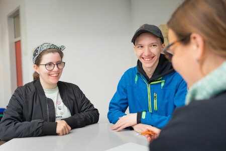 Zwei Personen stehen an einem weißen Tisch, eine dritte Person steht ihnen gegenüber und hält einen Stift in der Hand.