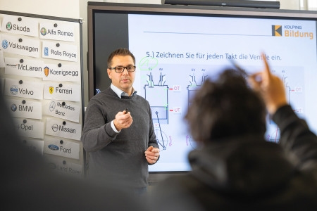 Lehrkraft steht vor einer Präsentation mit technischen Zeichnungen und Markenlogos von Automobilherstellern an der Wand, während eine Person im Publikum die Hand hebt.