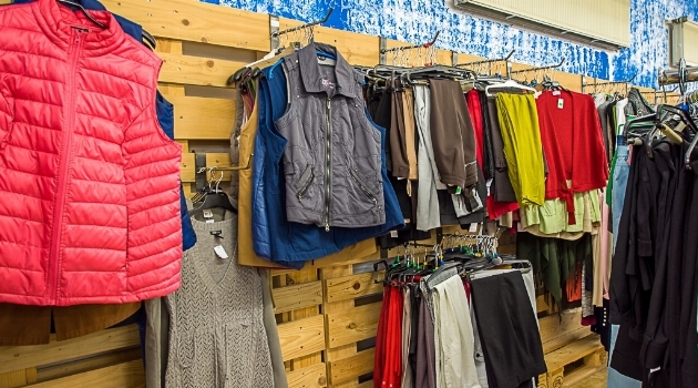 Mehrere Kleidungsstücke wie Jacken, Westen und Pullover hängen an einer Holzwand in einem Second-Hand-Laden.