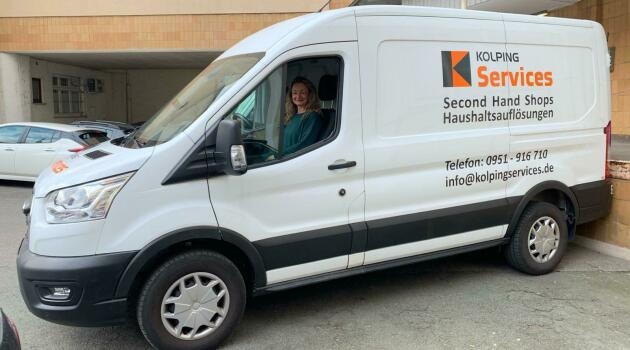 Weißer Transporter mit der Aufschrift 'KOLPING Services Second Hand Shops Haushaltsauflösungen' und Kontaktdaten, geparkt vor einem Gebäude.
