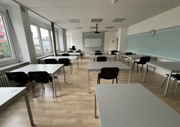 Leerer Seminarraum mit mehreren Tischen und Stühlen, großen Fenstern an der linken Seite, einer Tafel an der rechten Wand und einer Projektionsfläche vorne.
