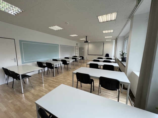 Leerer Seminarraum mit mehreren weißen Tischen und schwarzen Stühlen, großer grüner Tafel an der Wand und Projektionsleinwand am vorderen Ende.