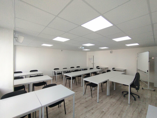 Leerer Schulungsraum mit mehreren Tischen und Stühlen, Deckenbeleuchtung und Whiteboard auf Rollen.