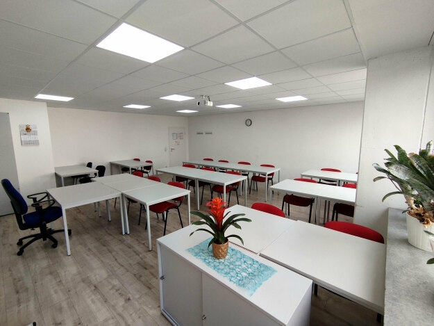 Schulungsraum mit mehreren weißen Tischen, roten Stühlen, einem Schreibtisch mit Pflanze und weißer Decke mit Leuchtpaneelen.