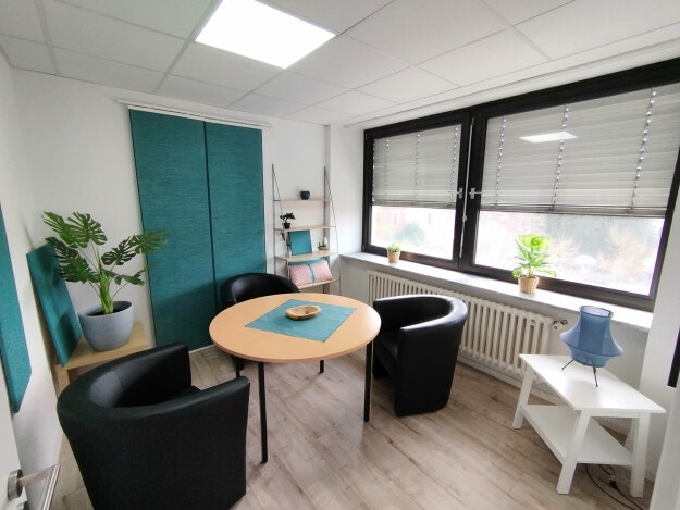 Kleiner Besprechungsraum mit rundem Holztisch, drei schwarzen Sesseln, zwei großen Fenstern mit halb geschlossenen Jalousien, Pflanzen auf Fensterbank und Möbeln sowie einem weißen Beistelltisch mit blauer Lampe.