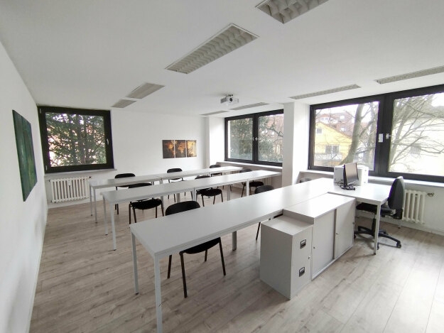 Heller Schulungsraum mit mehreren weißen Tischen, schwarzen Stühlen, großen Fenstern und einem Schreibtisch mit Computer.