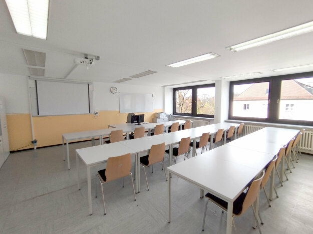Heller Schulungsraum mit drei langen Tischen, mehreren Holzstühlen und großen Fenstern an der rechten Seite.