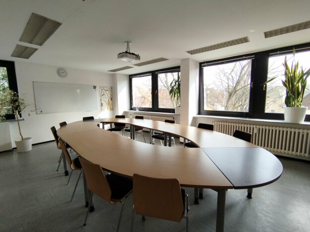 Konferenzraum mit ovalem Holztisch, mehreren Stühlen, großen Fenstern und einem Whiteboard an der Wand.