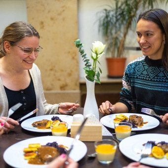 Zwei Personen sitzen an einem gedeckten Tisch mit Tellern voller Essen, Gläsern mit einer Nachspeise und einer weißen Vase mit einer weißen Blume in der Mitte.