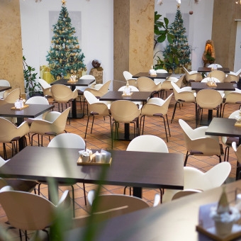 Leerer Gastraum mit mehreren Tischen und Stühlen vor einem geschmückten Weihnachtsbaum.