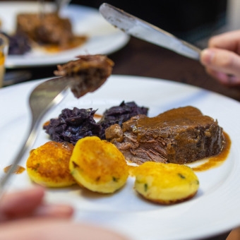 Nahaufnahme eines Tellers mit einem Stück Fleisch, Rotkohl und mehreren Kartoffelscheiben, eine Hand hält ein Messer und eine hält eine Gabel mit einem Stück Fleisch.