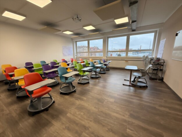 Klassenzimmer mit mehreren bunten Stühlen mit integrierten Schreibtischen vor großen Fenstern und Holzboden.