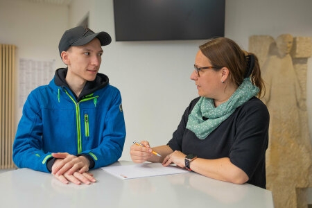 Ein junger Mann mit Basecap und eine Frau stehen an einem Tisch, eine hält einen Stift und ein Blatt Papier liegt vor ihnen.